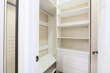 clean, white walk-in closet with shelves, drawers, and a hanging rod, partially visible from an open doorway