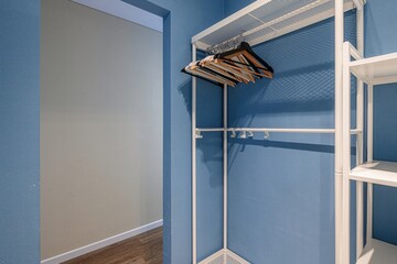 close shot features a white, minimalist open wardrobe with clothes hangers against a blue wall in a...