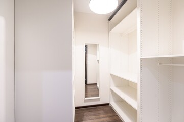 bright walk-in closet with white shelves, a full-length mirror, and dark wood floors. Clean, modern design with ample storage space