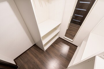 Obraz premium well-lit walk-in closet features white shelving units against a white wall, complemented by a dark wood floor and a mirror reflecting a modern door