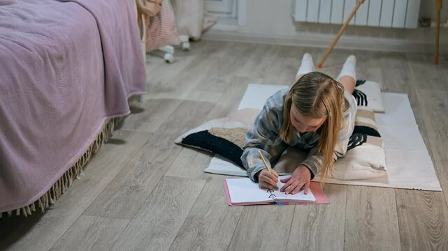 Caucasian teenage girl is thinking and writing in copybook with pen lying on the floor in bedroom. Female kid is engaged in home education solving mathematics problems and taking notes. Learning theme