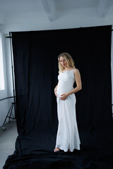 Beautiful pregnant young blonde in a long white silk dress to the floor on a black fabric...