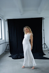Beautiful pregnant young blonde in a long white silk dress to the floor on a black fabric...
