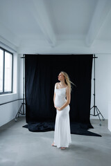 Beautiful pregnant young blonde in a long white silk dress to the floor on a black fabric...