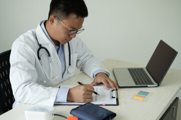 Young asian male doctor with stethoscope is busy with phone call while taking notes, using laptop,...