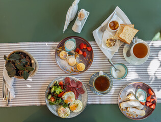 Overhead view of assorted breakfast plates with eggs syrniki cottage cheese rolls strawberries and hot black tea