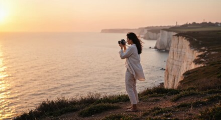 Woman photographing sunset on coastal cliff with serene ocean view