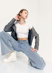 A stylish woman in trendy jeans and a jacket poses sitting on the floor