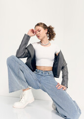 A stylish woman in trendy jeans and a jacket poses sitting on the floor