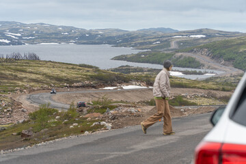 A woman in a gray beanie and beige jacket crosses a winding road surrounded by the Arctic tundra...