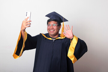 Young man dressed in cap and gown to celebrate his graduation uses his cell phone to take selfies,...
