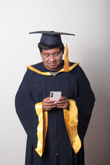Young man dressed in cap and gown to celebrate his graduation uses his cell phone to take selfies,...