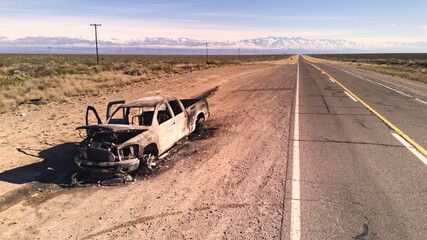 Burnt remains of a pickup truck by an asphalt road in the desert. Aerial view. No AI.