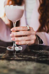 Elegant woman's hand with manicured nails holding a pink creamy drink in a glass, resting on a marble table, adorned with pearl jewelry. Close-up, stylish and refined.
