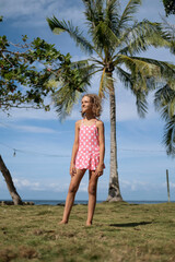 Wearing a trendy coral swimsuit, a teenage girl stands confidently against palm trees and a vast...