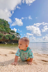 Jogja, indonesia-January 13, 2023. Asian toddler girl crawling happily on sandy beach with sea and hills behind. Fun childhood, tropical nature, family vacation, outdoor play, and carefree summer day.
