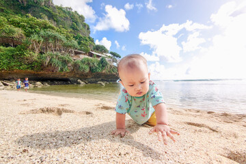 Jogja, indonesia-January 13, 2023. Asian toddler girl crawling happily on sandy beach with sea and hills behind. Fun childhood, tropical nature, family vacation, outdoor play, and carefree summer day.