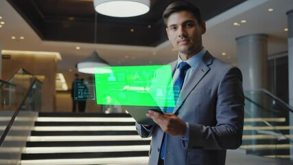 A young South Asian man in a suit holds a tablet displaying a green screen interface. The background features a modern interior with stairs and lighting. - Powered by Adobe