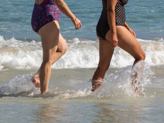 Fototapeta premium Mature women walk barefoot on the beach, wading into the Mediterranean Sea, foam waves lapping at their legs. Scenic portrait overlooking the coast. Majorca, or Mallorca. Balearic Islands, Spain.