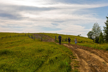Family walking on mountain trail at sunset. Three people walk along a dirt path in the mountains,...