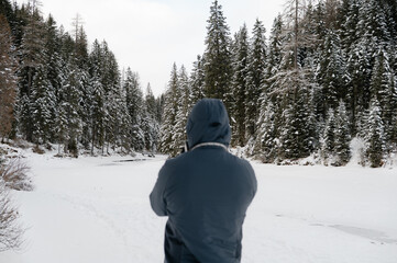 A man in a blue jacket is standing in the snow, taking a picture of the forest
