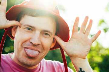 A man playfully poses outdoors in a bright red hat, sticking out his tongue in the midst of...
