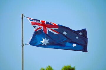 The Australian flag flies proudly up a flagpole on a beautiful summer day with a background of blue...