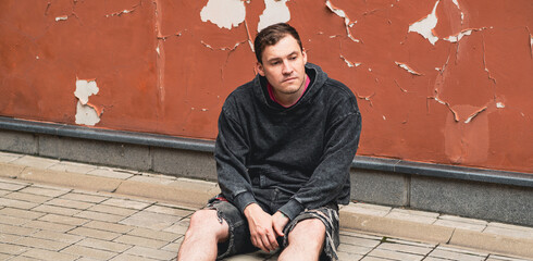 A man wearing a gray hoodie appears lost in thought as he sits against a faded orange wall The wall...