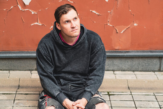 A man wearing a gray hoodie appears lost in thought as he sits against a faded orange wall The wall showcases peeling paint creating a textured background in the afternoon light - Powered by Adobe