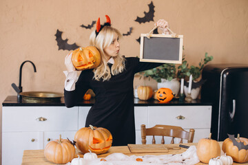 Woman wearing witch costume holding carved pumpkin and empty blackboard in Halloween decorated...