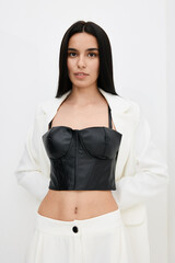 young woman wearing black leather bustier and white blazer, posing confidently against plain white...