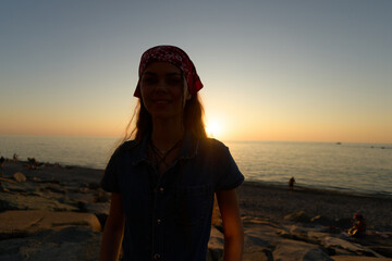 woman standing at the beach during sunset wearing a red headscarf and denim clothing with calm sea...