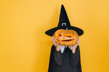 Little witch holding jack o' lantern in front of her face posing on yellow background