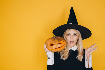 Surprised witch holding jack o' lantern posing on yellow background