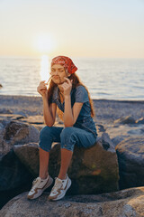 young woman sitting on rocks by the sea wearing a red headscarf and denim overalls at sunset with a...