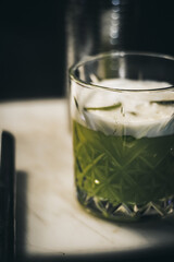 A foamy green cocktail in a crystal glass on a white marble surface with soft focus and dark background.
