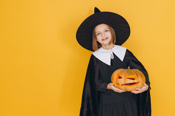 Smiling girl wearing witch costume holding jack o' lantern posing on yellow background