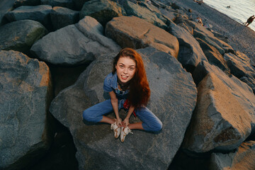 Lifestyle woman in modern boho-western denim street style sitting on rocks by the shore, with warm...