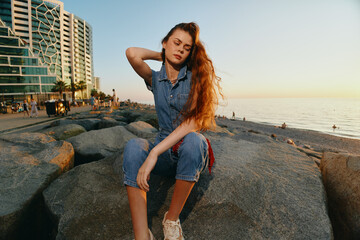 relaxed woman sitting on rocks by the sea at sunset wearing denim outfit with long red hair and...