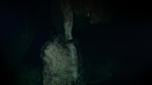 Sunlight barely pierces through the cavern's entrance, illuminating jagged rocks. The darkness creates an eerie atmosphere as the deep blue water swallows the hidden wonders of Yucatan.
