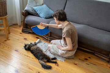 Tired sweaty woman fans cat to provide relief at home without AC. Pet care under high temperatures,...