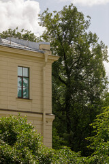 Elegant corner of a classic beige building with a single window, surrounded by lush green trees and a partly cloudy sky.