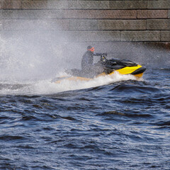 A person riding a yellow jet ski creates a splash as they speed across the water near the stone embankment.