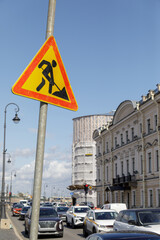 A construction warning sign stands next to a busy street in Saint Petersburg, with cars passing and a building under renovation in the background.