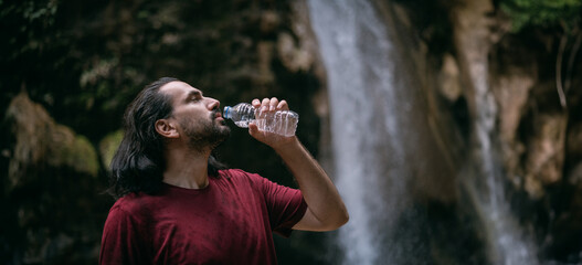 A young man drinks water from a bottle near a tropical waterfall among the rocks.
