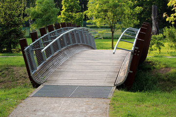 A uniquely arched wooden footbridge with sleek metal railings spans a grassy park, blending contemporary design with natural surroundings and inviting pedestrian passage