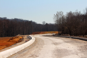 A newly constructed, curving road with concrete barriers winds through a forested area, bordered by...