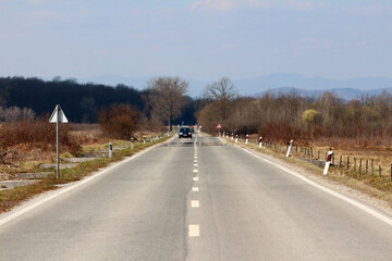 A straight, two-lane rural road stretches into the distance, bordered by dry grass and shrubs, with a single car approaching and white roadside markers lining both sides