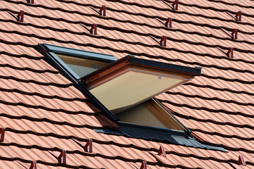 A contemporary roof window is propped open on a sloped roof covered with red terracotta tiles,...