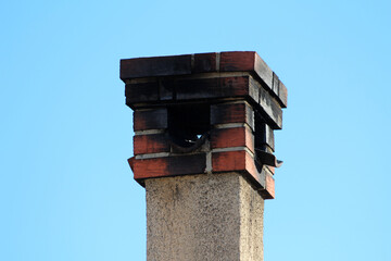 A weathered brick chimney with a blackened, soot-stained top rises against a clear blue sky,...